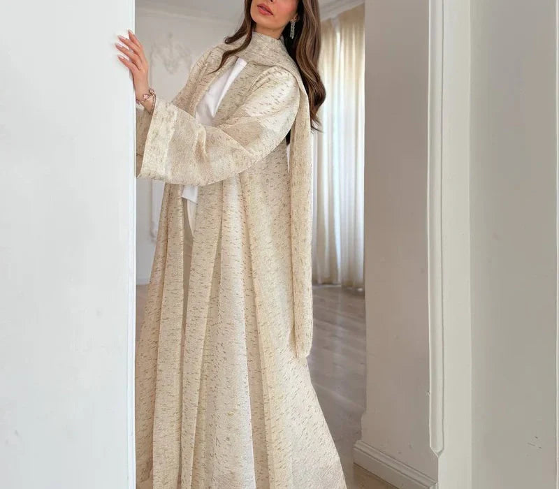 A woman wearing a light beige abaya stands gracefully near a doorway in a softly lit interior.